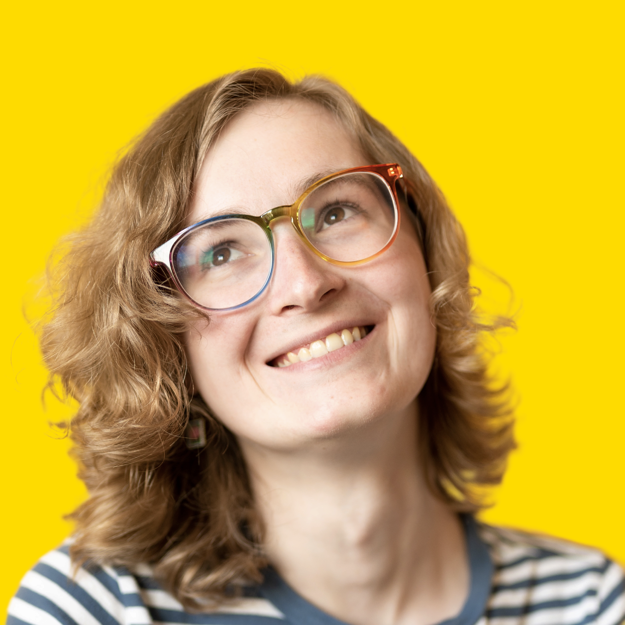 A picture of Claire Violet with a very big and genuine smile, looking up and away from the camera. Claire is wearing rainbow glasses and has somewhat curly blonde shoulder-length hair. In the background is a wall painted a bright saturated yellow color.